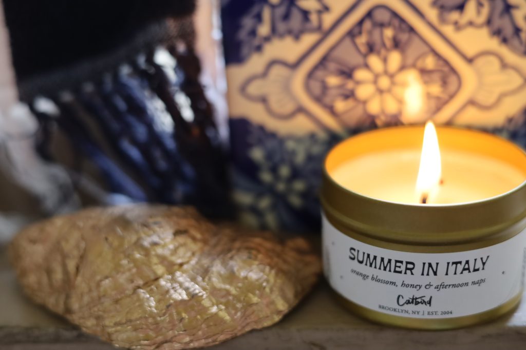 summer in italy candle burning with an italian blue and white tile in the background, a beach towel and oyster shell